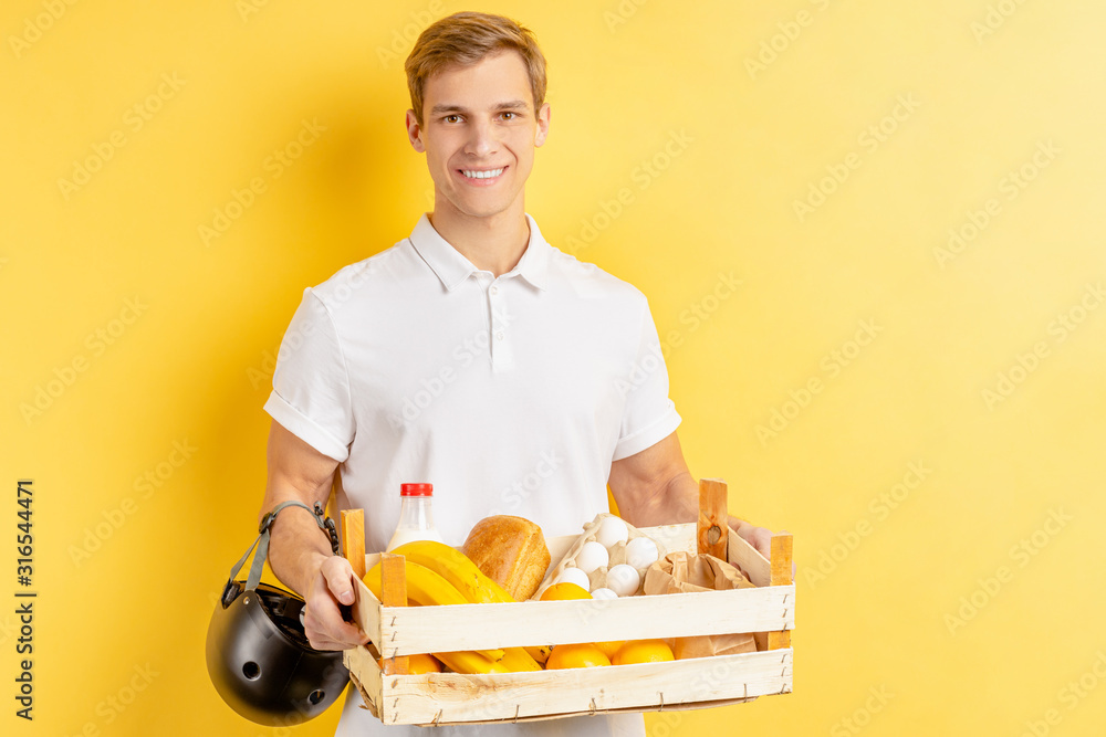 portrait of smiling young delivery man holding fresh vegetables and fruits in box, look at camera. isolated over yellow background portrait of smiling young delivery man holding fresh vegetables and fruits in box, look at camera. isolated over yellow background