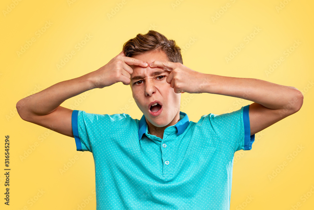 Cosmetology, Dermatology and acne. Caucasian teenage boy in a blue t-shirt, painfully squeezes out a pimple on his forehead. Yellow background. Copy space Cosmetology, Dermatology and acne. Caucasian teenage boy in a blue t-shirt, painfully squeezes out a pimple on his forehead. Yellow background. Copy space