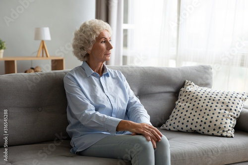 Thoughtful older woman sitting on couch, lost in thoughts