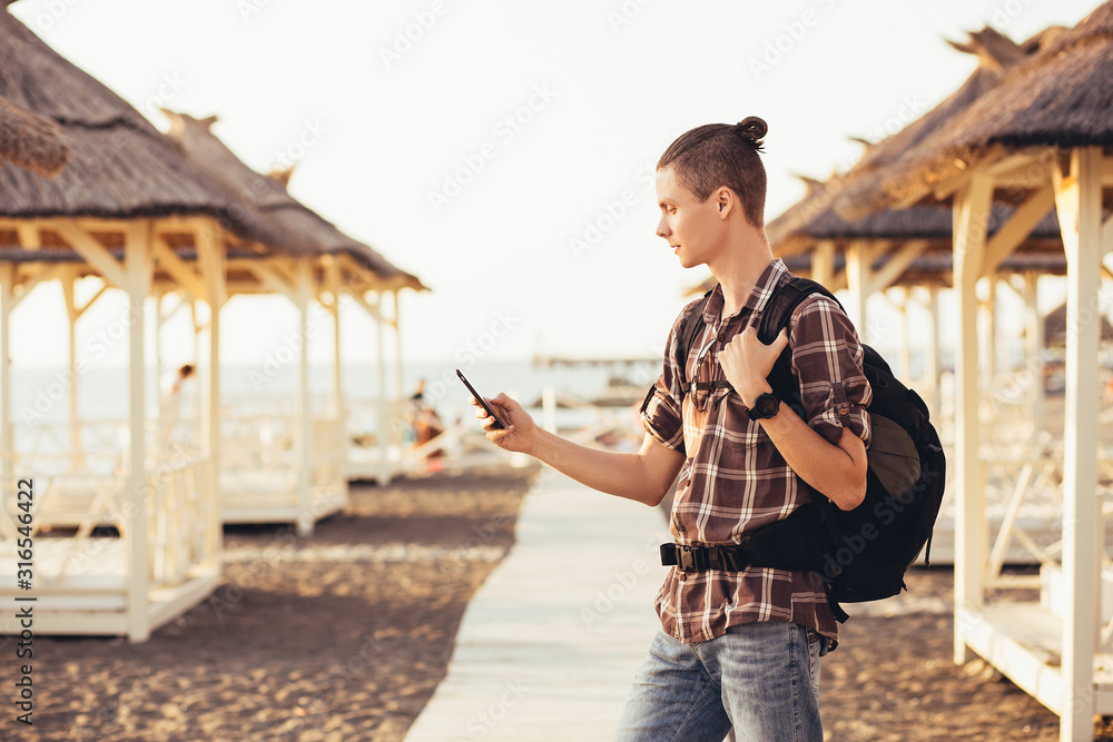 Side view - a young traveler guy with a backpack is looking at a smartphone and is guided around the map in search of a hotel. The concept of online navigators in the smartphone