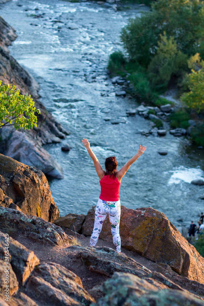 adult woman doing yoga asanas on the rocks above the white water river at sunset time