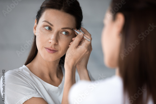 Beautiful woman tweezing eyebrows close up, looking in mirror