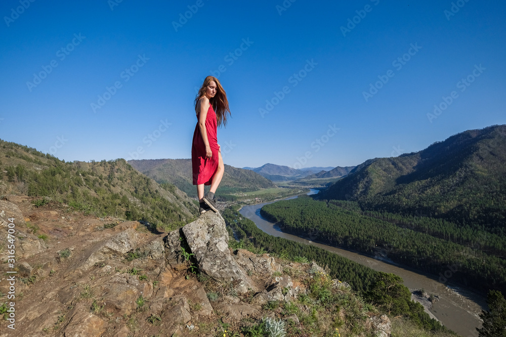 Naklejka premium Portrait of a woman in a red dress on top of a mountain on the background of the river flowing between the rocks.