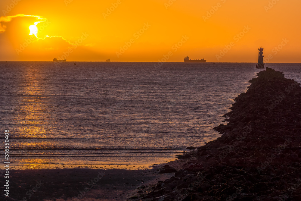 Silhouette of cargo ships at golden hour. Sun, solar disc, shines from behind cloud. Lighthouse and breakwater on Bull Island, Dublin, Ireland Silhouette of cargo ships at golden hour. Sun, solar disc, shines from behind cloud. Lighthouse and breakwater on Bull Island, Dublin, Ireland
