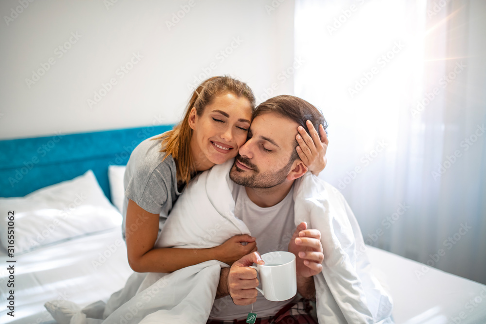 Caring wife. Young woman taking care of his sick husband, bringing tea to bed. Couple with cold in bed at home together. Nice caring woman touching her husbands forehead while sitting near him