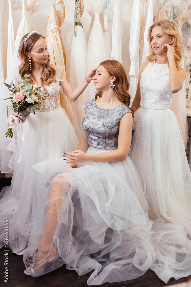 portrait of three attractive young ladies wearing fashionable airy dresses stand together in wedding salon, posing at camera. try on new dress by professional dress designer
