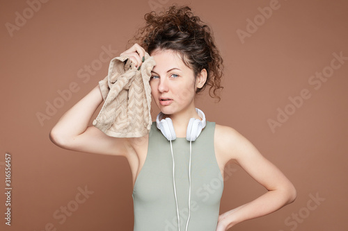 Sensual fitness woman in top and headphones on neck takes break after training, wipes sweat with towel, being energetic runner or jogger, feels tired, but confident, lookingwith blue eyes at camera.