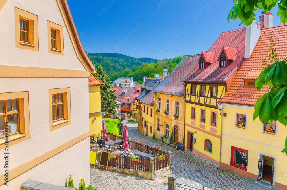 Loket town narrow street, colorful traditional typical buildings with multicolored walls, cobblestone road, wooden fence, green hills background, Karlovy Vary Region, West Bohemia, Czech Republic