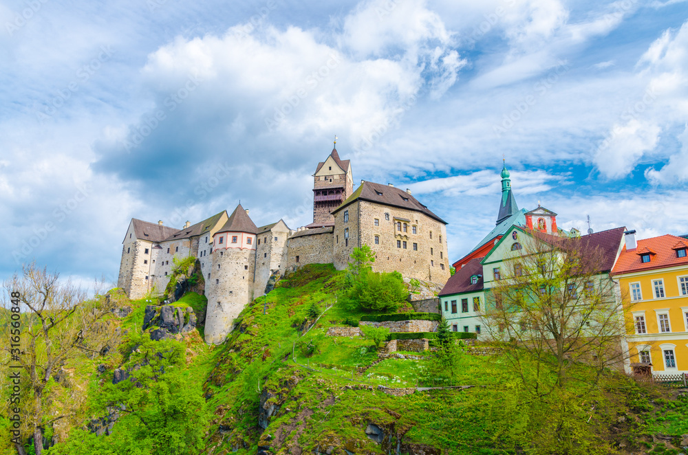 Loket Castle Hrad Loket gothic style building on massive rock and colorful buildings in Loket town, green trees and hills, blue cloudy sky background, Karlovy Vary Region, West Bohemia, Czech Republic