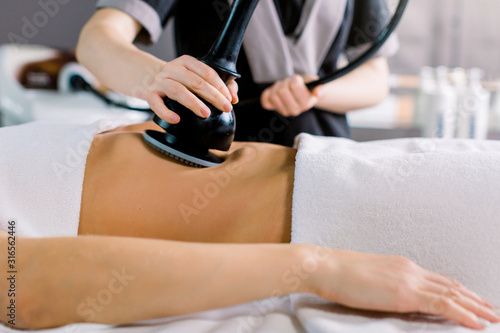 Cropped shot of hands of cosmetologist doing ultrasound cavitation procedure on the belly of woman. Unrecognizable woman getting anti-fat treatment at beauty salon. Abdominal slimming concept