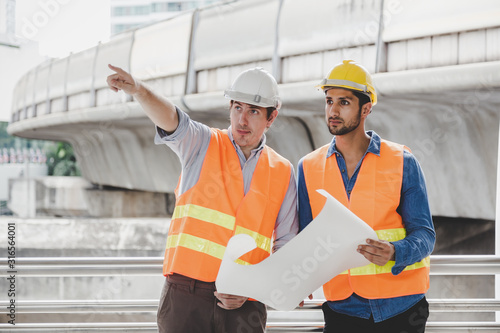 Engineer boss and foreman or worker working together at worksite in city. Employer guy pointing finger to construction for explaining project or plan. Blue collar worker hold blueprint, safety hats
