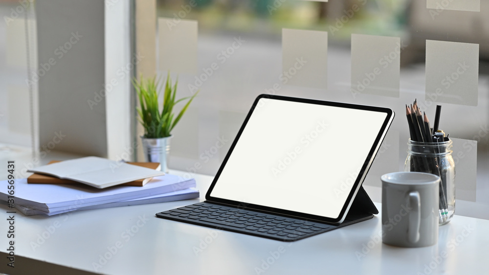 Photo of white blank screen computer tablet in a leather-case and keyboard is setting on a modern working table over comfortable office background. Photo of white blank screen computer tablet in a leather-case and keyboard is setting on a modern working table over comfortable office background.