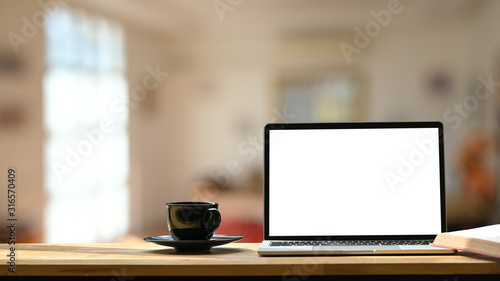 Photo of white blank screen laptop, black coffee cup and coasters on the wooden working desk over blurred modern cafe background.