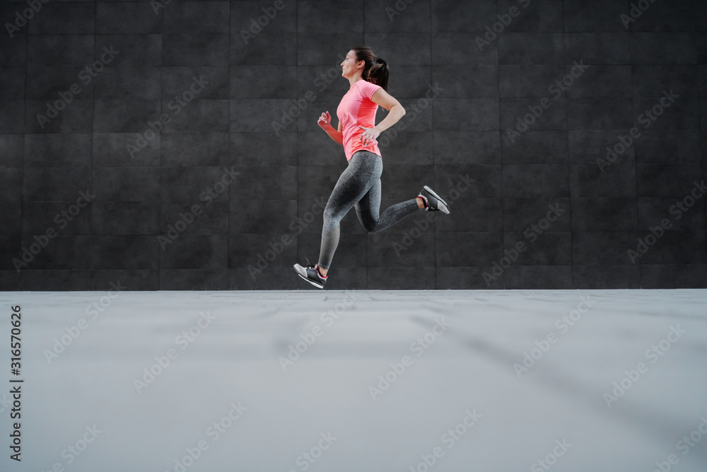 Side view of fit attractive Caucasian woman in sportswear and with ponytail running outdoors. In background is dark wall. Side view of fit attractive Caucasian woman in sportswear and with ponytail running outdoors. In background is dark wall.