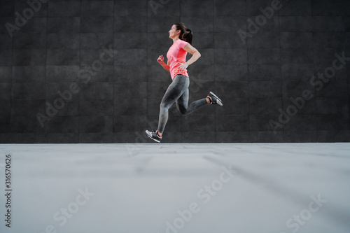 Side view of fit attractive Caucasian woman in sportswear and with ponytail running outdoors. In background is dark wall. © dusanpetkovic1