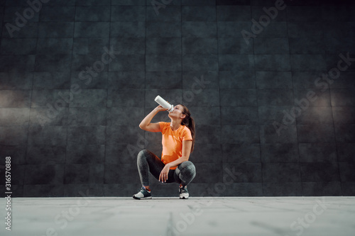Charming fit caucasian sportswoman in shape dressed in active wear crouching and drinking water. In background is dark wall. © dusanpetkovic1