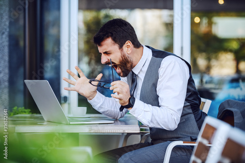 Obraz na plátně Angry elegant businessman in suit looking at laptop and yelling while sitting in cafe