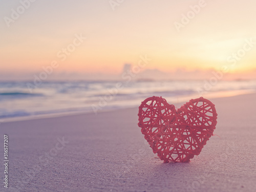 Tropical background for St. Valentine's Day. A red wicker heart is located on a sandy beach. Behind is the sea or ocean. Love of travel.