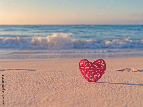 Tropical background for St. Valentine's Day. A red wicker heart is located on a sandy beach. Behind is the sea or ocean. Love of travel.