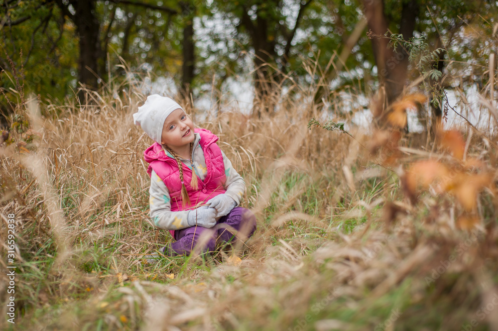 Little beautiful girl in a hat, in warm clothes and gloves sits on the dry grass in the autumn park. Little beautiful girl in a hat, in warm clothes and gloves sits on the dry grass in the autumn park.