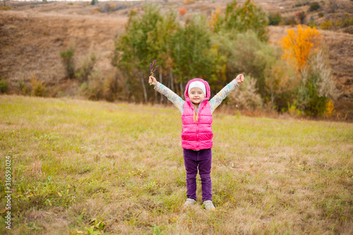 Little beautiful girl stands in a field holding her hands up on a background of autumn trees.