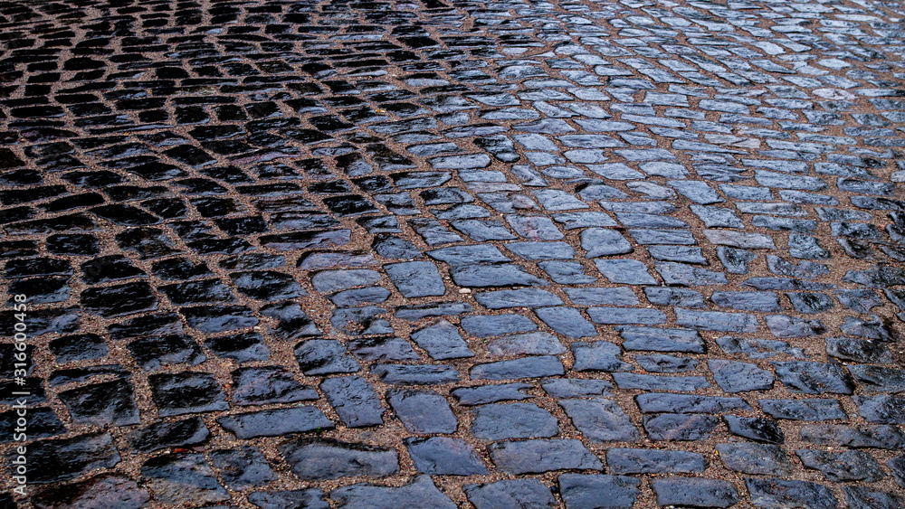 The old, wet pavement paved with large cobblestones on a sandy base in the evening in cloudy weather is shiny after the rain. Natural background from the road surface. The old, wet pavement paved with large cobblestones on a sandy base in the evening in cloudy weather is shiny after the rain. Natural background from the road surface.