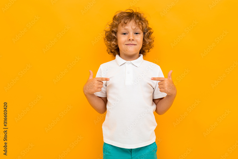 Little boy with curly hair in colourful t-shirt and shorts shows thumbs for himself isolated on yellow background - obrazy, fototapety, plakaty 