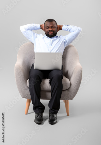African-American man with laptop sitting in armchair against grey background