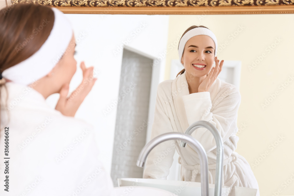 Beautiful young woman applying cream on her face near mirror in bathroom Beautiful young woman applying cream on her face near mirror in bathroom