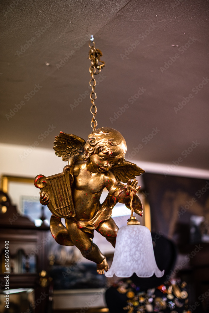 Foto Stock Statue of angel. Gold angel. A flower arrangement hanging from  ceiling with lamp wooden background. On chain old vintage house items sale  storage container uk text advertisement | Adobe Stock
