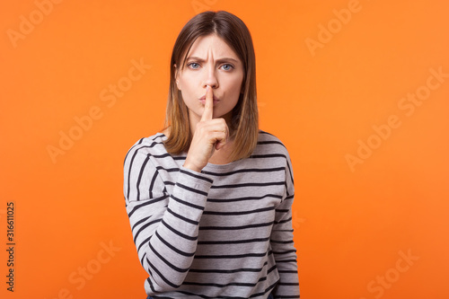 Be quiet! Portrait of serious woman with brown hair in long sleeve striped shirt standing, making silence gesture, looking with frowning worried face. indoor studio shot isolated on orange background