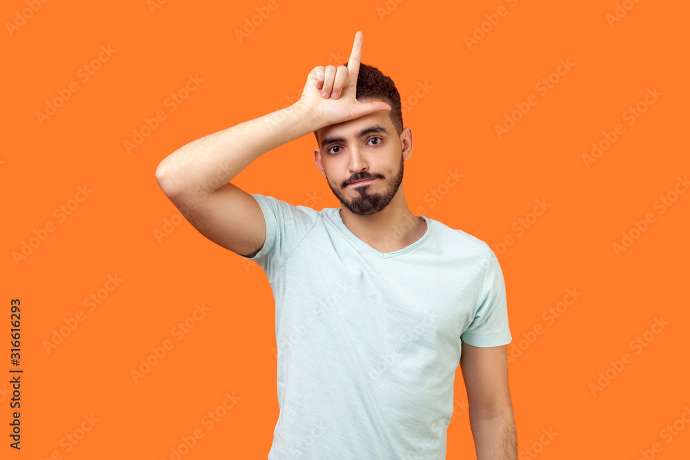 I am loser! Portrait of unlucky brunette man with beard in casual white t-shirt standing with grumpy face and showing loser gesture, upset over defeat. indoor studio shot isolated on orange background I am loser! Portrait of unlucky brunette man with beard in casual white t-shirt standing with grumpy face and showing loser gesture, upset over defeat. indoor studio shot isolated on orange background