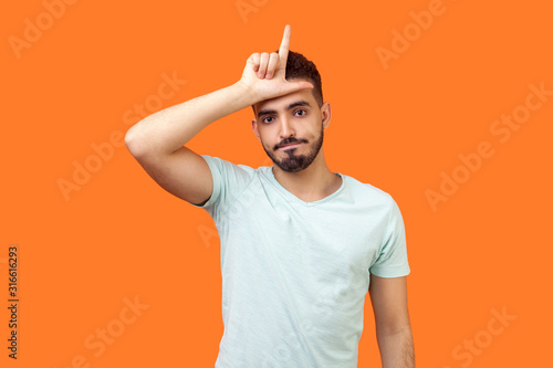 Photos I am loser! Portrait of unlucky brunette man with beard in casual white t-shirt standing with grumpy face and showing loser gesture, upset over defeat
