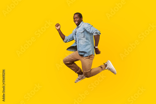 Full length portrait of happy joyous man in denim casual shirt jumping or flying  hurry running to his dream  looking at camera with toothy smile. indoor studio shot isolated on yellow background