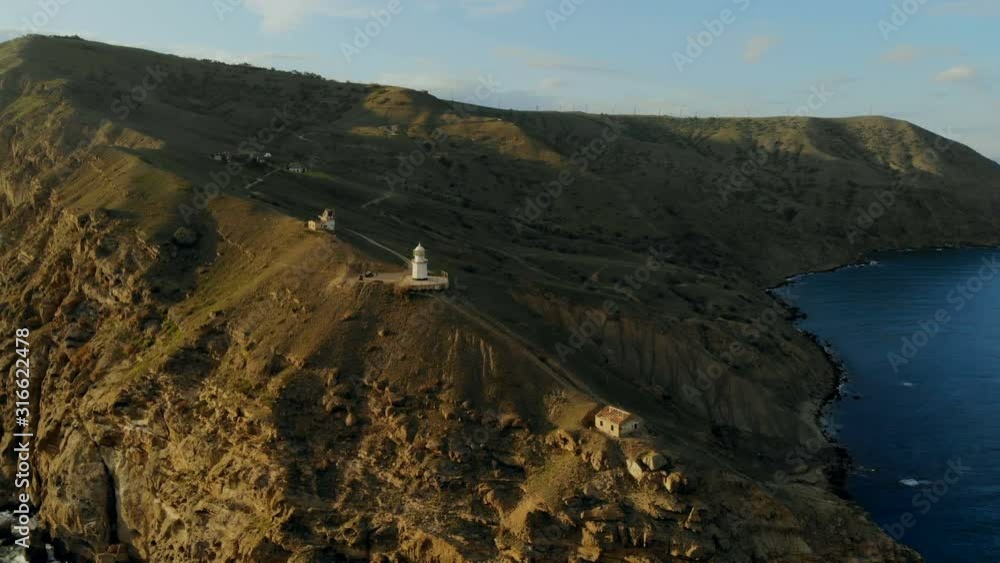 Drone flight at sunset over the lighthouse, surrounded by turquoise green sea and mountain slopes Drone flight at sunset over the lighthouse, surrounded by turquoise green sea and mountain slopes