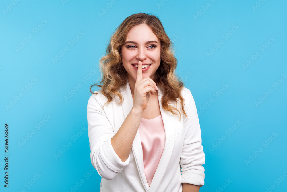 Please, be quiet! Portrait of positive lady with wavy hair in white jacket smiling and making hush sign with finger on lips, asking for silence and secrecy, keep calm. studio shot, blue background Please, be quiet! Portrait of positive lady with wavy hair in white jacket smiling and making hush sign with finger on lips, asking for silence and secrecy, keep calm. studio shot, blue background