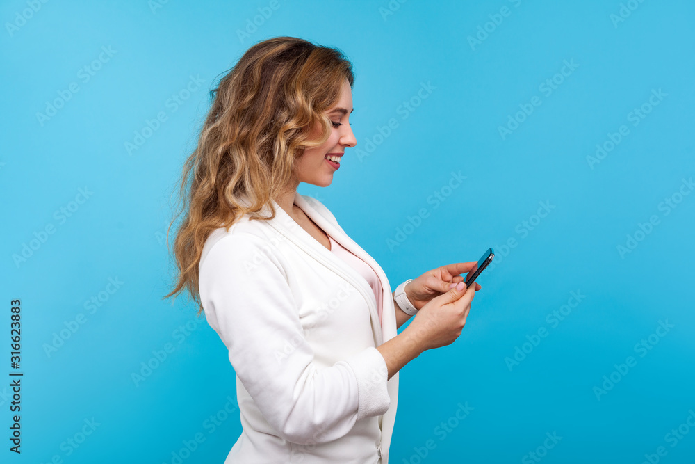 Side view of positive charming woman with wavy hair in white jacket typing message on cellphone and smiling, using mobile apps, online communication in social network. studio shot, blue background Side view of positive charming woman with wavy hair in white jacket typing message on cellphone and smiling, using mobile apps, online communication in social network. studio shot, blue background
