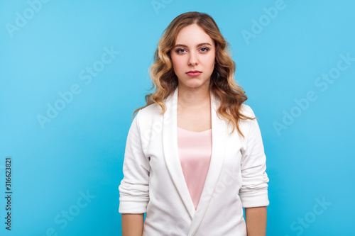 Portrait of serious upset unsmiling beautiful woman in casual white jacket standing looking at camera with sad eyes, unhappy determined face expression. indoor studio shot isolated on blue background © khosrork