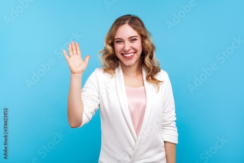 Hello! Portrait of friendly kind cheerful girl with wavy hair in white jacket waving hand saying hi welcome, smiling with hospitable sociable expression. indoor studio shot isolated on blue background photo