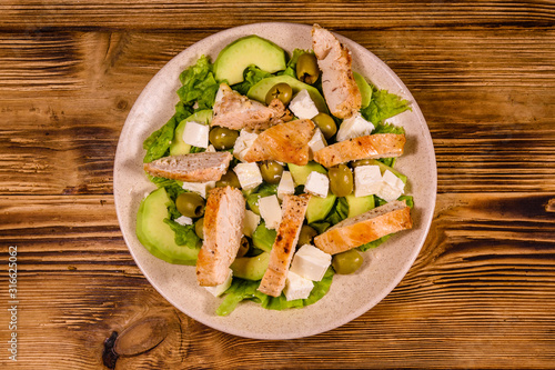 Fresh salad with chicken meat, feta cheese, avocado, green olives and lettuce leaves in ceramic plate on wooden table. Top view