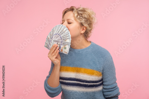 Portrait of pleased happy woman with curly hair in warm sweater smelling dollar banknotes and closing eyes from pleasure and satisfaction, greed for money. studio shot isolated on pink background photo