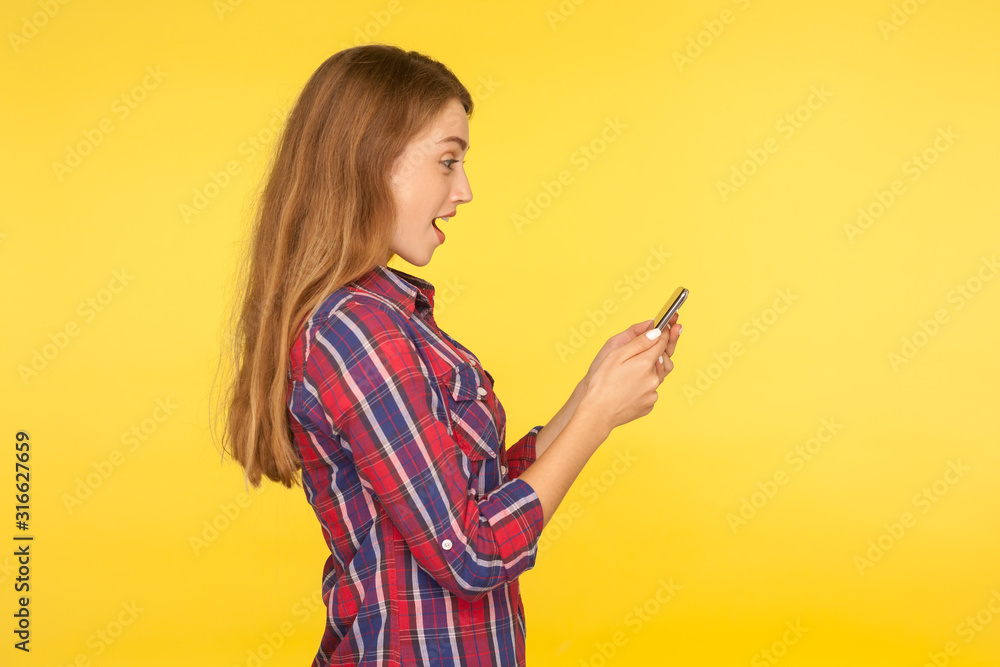 Side view of amazed ginger girl in casual shirt using cell phone and looking shocked pleasantly surprised by online mobile services, typing message. indoor studio shot isolated on yellow background Side view of amazed ginger girl in casual shirt using cell phone and looking shocked pleasantly surprised by online mobile services, typing message. indoor studio shot isolated on yellow background