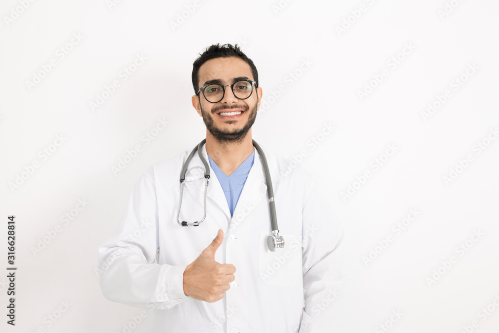 Young smiling clinician in eyeglasses and whitecoat showing thumb up