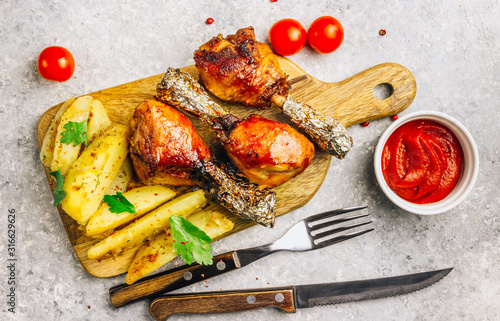Grilled chicken drumstick bbq with potato slices on a cutting board on a stone gray background.