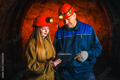 A beautiful young girl in a red helmet and with a electronic tablet in her hands is standing with a miner in a coal mine. Business plan discussion.