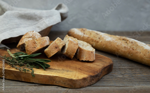 A whole-grain chopped baguette with herbs lies on a wooden board, next to a fresh sprig of thyme and rosemary herb. In the background a baguette and a wicker basket with bread on a wooden table