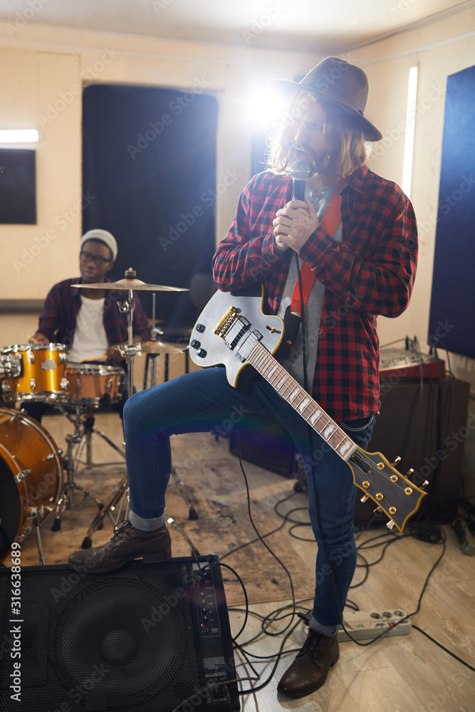 Obraz premium Full length portrait of contemporary long-haired man singing to microphone during rehearsal or concert with music band in background