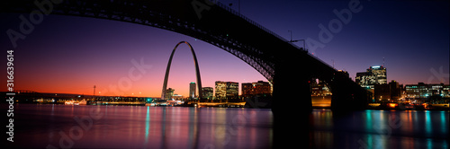 This is the skyline and Arch at sunset. Above it is the Eads Bridge along the Mississippi River. There is a purple cast in the sunset sky. photo