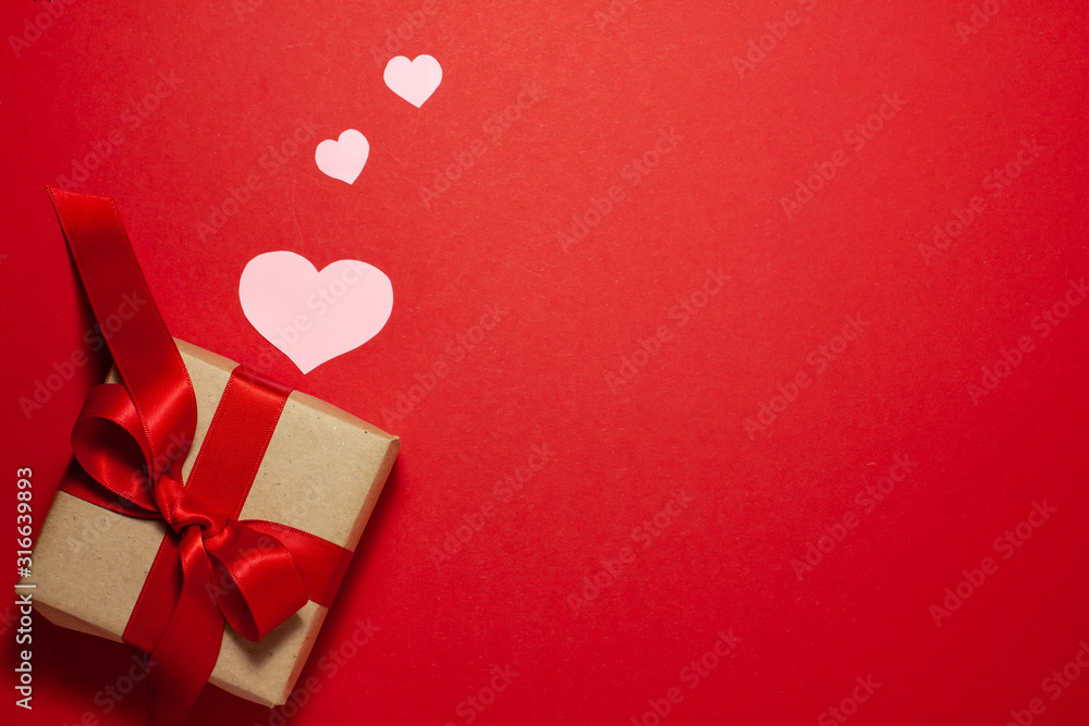 Pink hearts of different sizes near the brown paper gift box with a red ribbon bow on the left side of the frame. Valentines day concept. Flat lay top view with space for text on red background