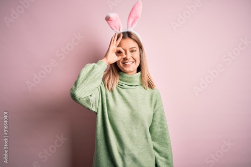 Photography Young beautiful woman wearing easter rabbit ears standing over isolated pink background doing ok gesture with hand smiling, eye looking through fingers with happy face